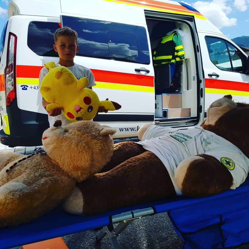 Kind mit einem Pikachu-Stofftier neben einem Krankenwagen und Kuscheltieren auf der Trage.