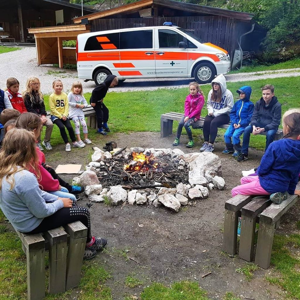 Kinder sitzen um ein Lagerfeuer, im Hintergrund ein Krankenwagen des Grünen Kreuzes.