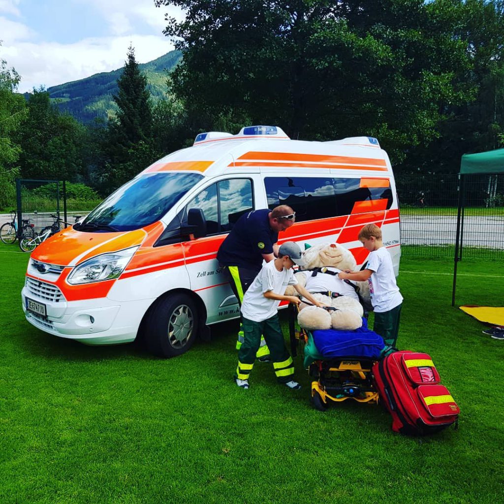 Kinder lernen Erste Hilfe an einem Rettungswagen mit einem großen Stoffbären.