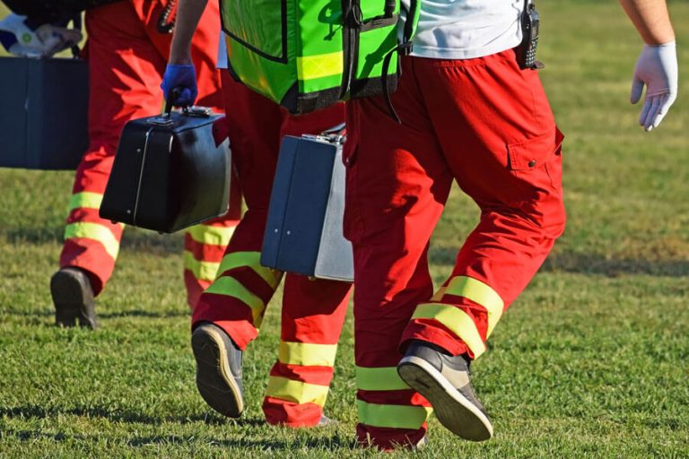 Spasilačke službe u crveno-žutim uniformama nose medicinsku opremu na livadi.