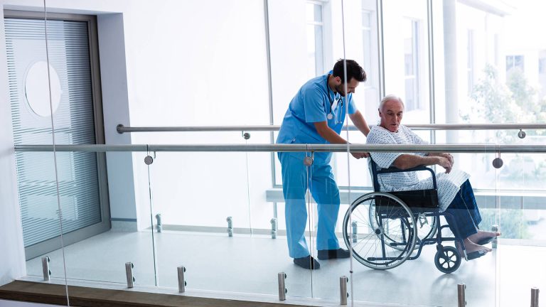 Njegovatelj gura pacijenta u invalidskim kolicima kroz moderan hodnik u zdravstvenoj ustanovi.