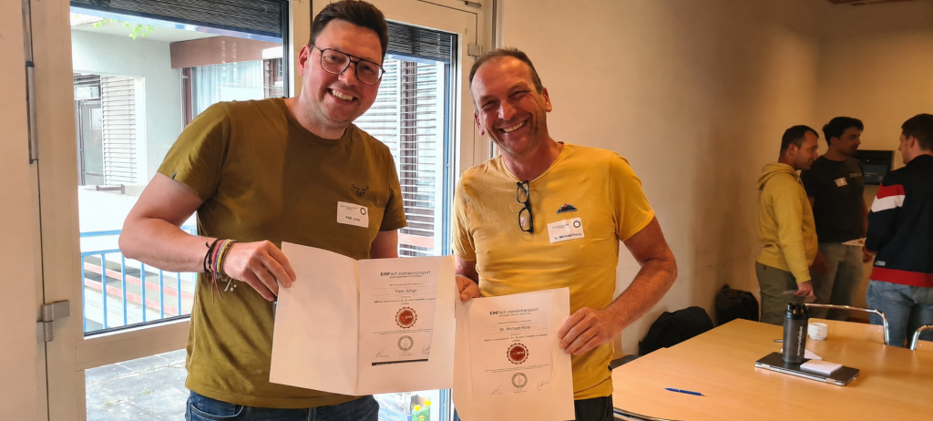 To smilende mænd holder certifikater i et undervisningslokale ved siden af store vinduer